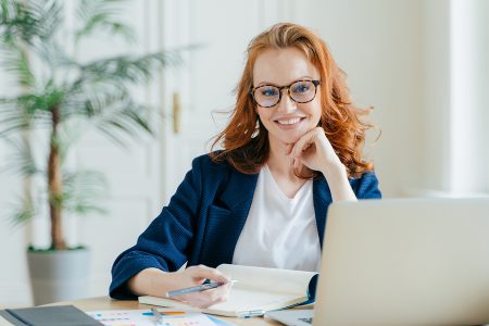 Photo of successful ginger female freelancer has remote work, watches webinar online on laptop computer, writes down information in notepad, smiles happily poses at workplace dressed in formal clothes