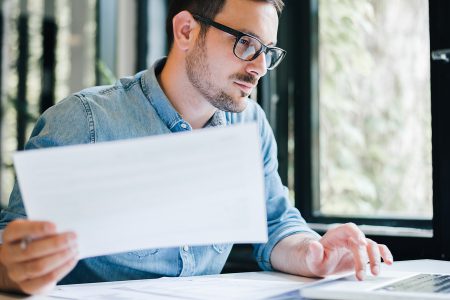 Serious pensive thoughtful focused young casual business accountant bookkeeper in office looking at and working with laptop and income tax return papers and documents