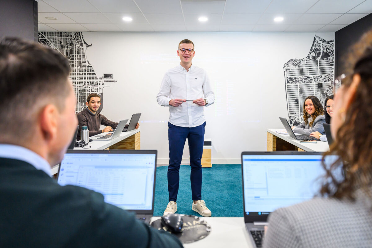 réunion de plusieurs personnes dans une salle, une personne est debout et parle au milieu. Il est entouré de ses collègues assis sur les ordinateurs qui l'écoutent